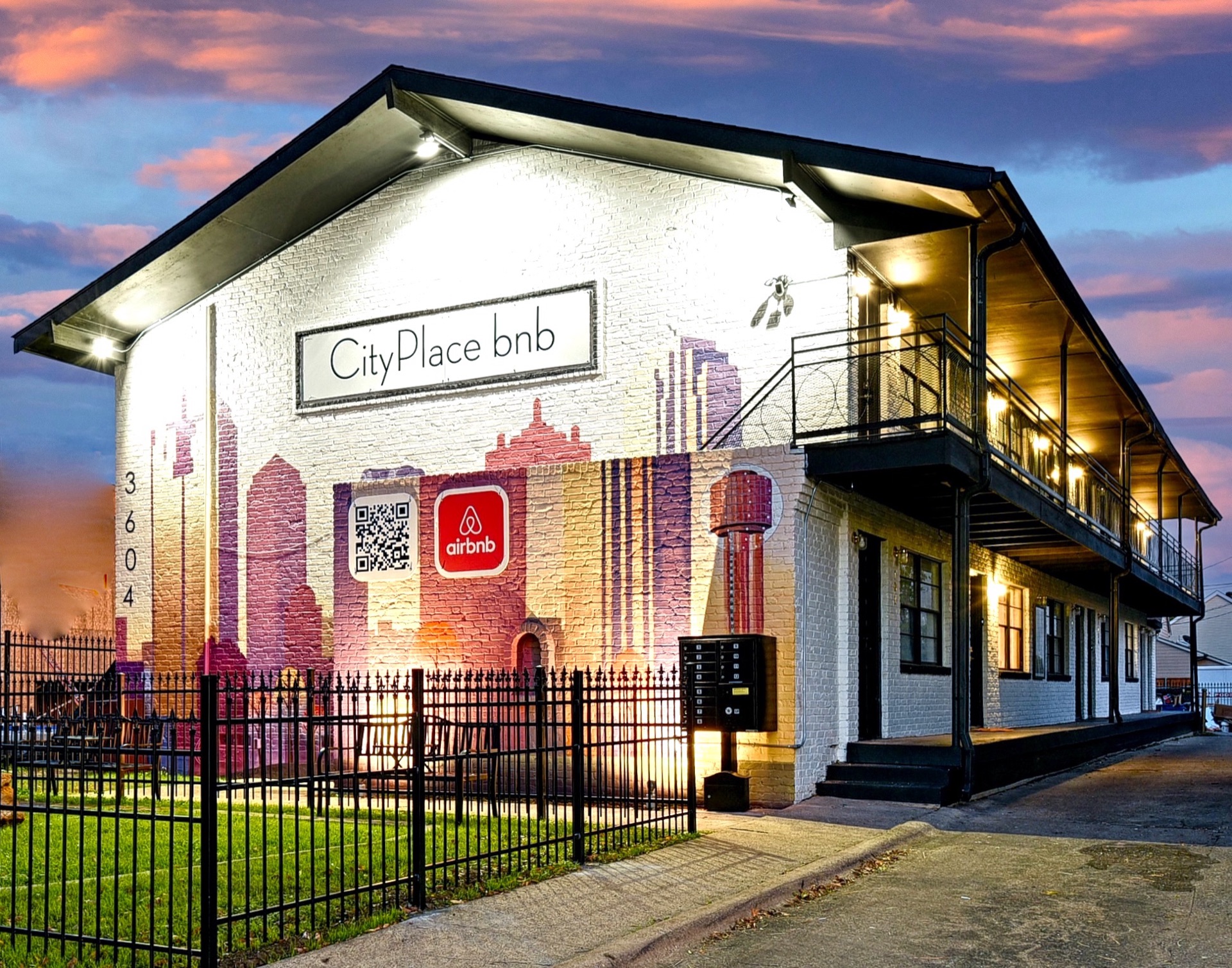 CityPlace bnb at dusk, two-story building with a Dallas-skyline mural across the facade, lawn and iron fence in front