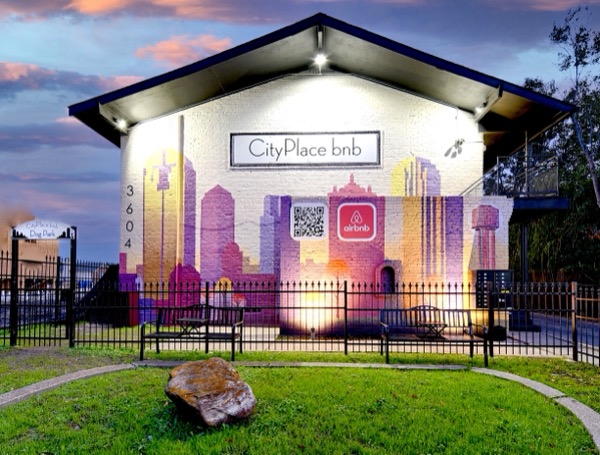 Dallas-skyline mural on the building gable above the fenced dog park lawn at dusk